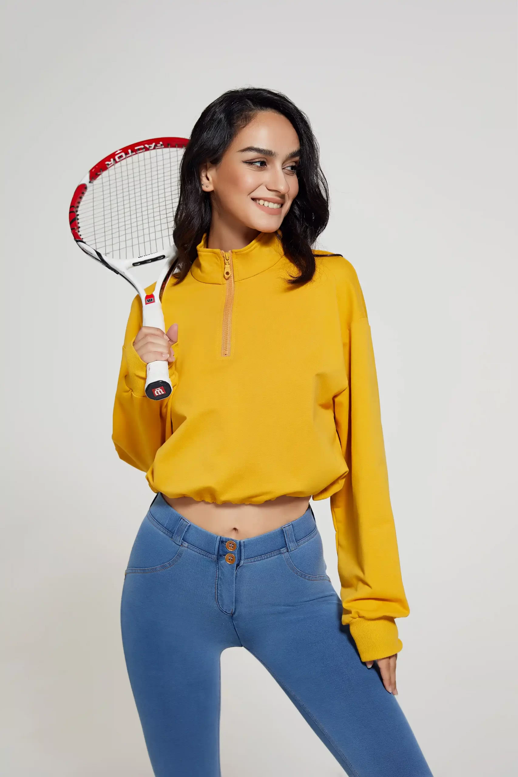 Female model in yellow cropped activewear top and high-rise blue leggings holding a tennis racket, showcasing sporty lifestyle wear